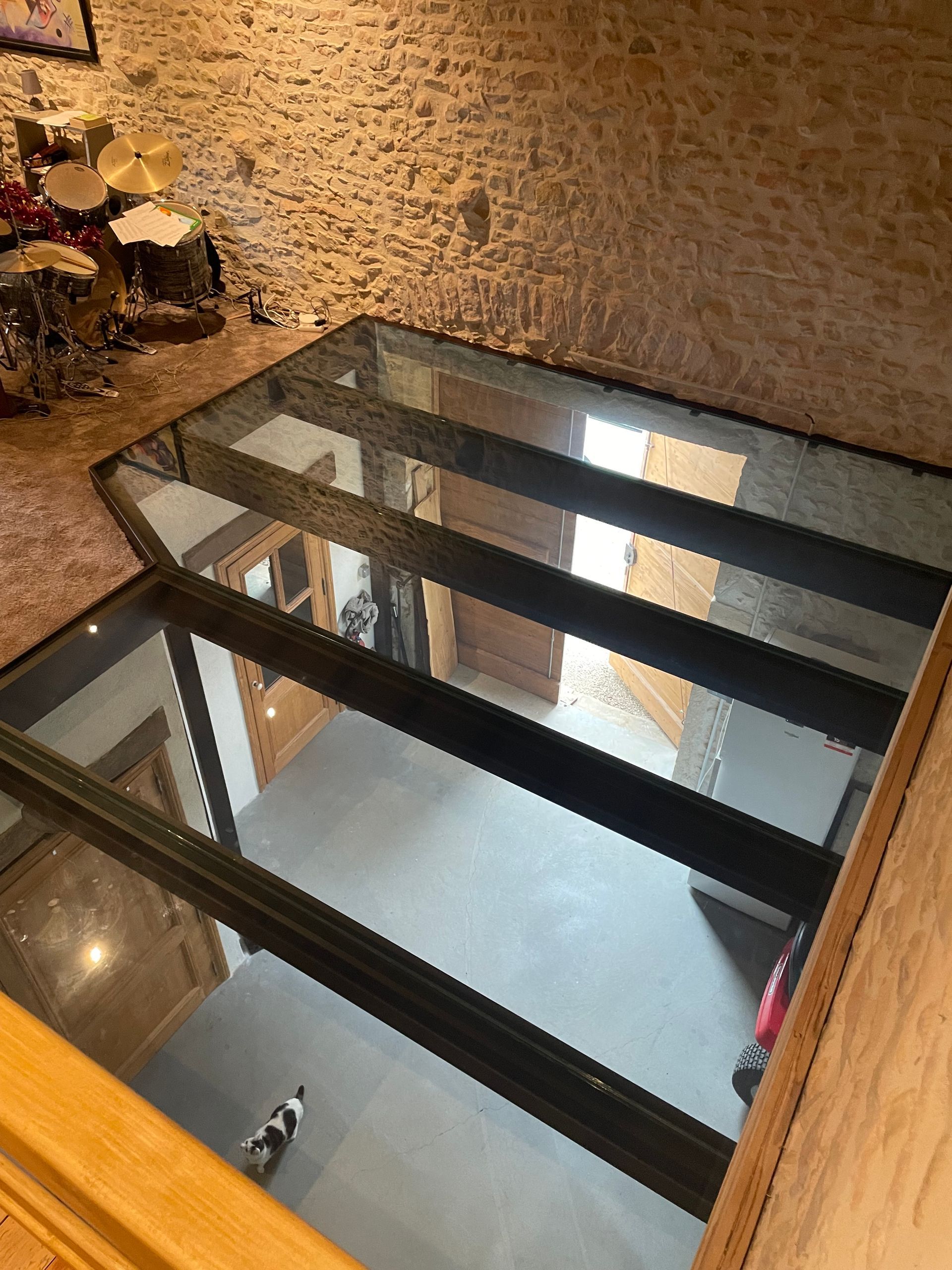 Dans une pièce rustique aux murs de pierre, une section de plancher en verre surplombe un niveau inférieur au sol gris où se trouve un petit chien.