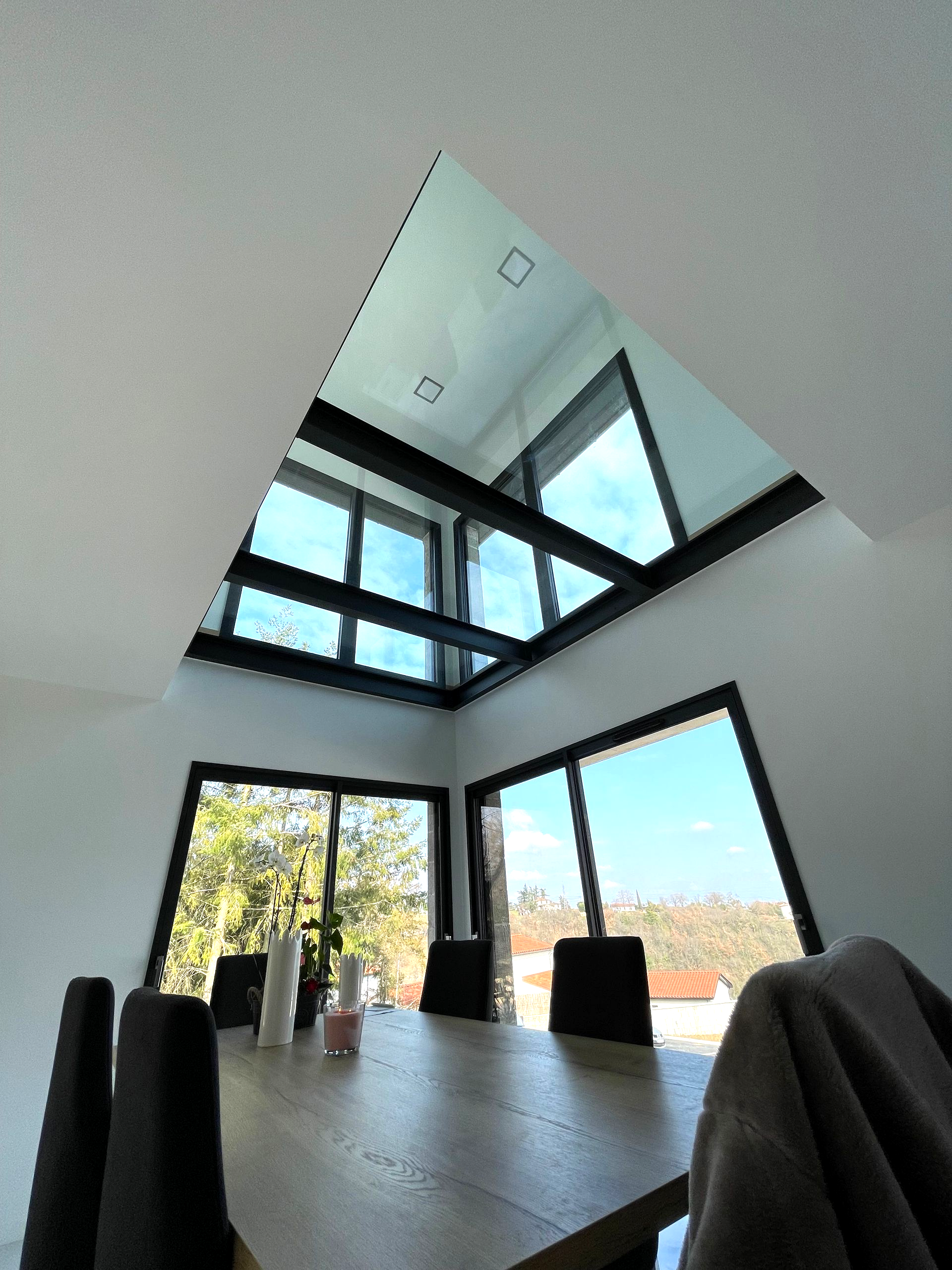 Salle à manger moderne avec une verrière triangulaire et de grandes fenêtres. On aperçoit une table et des chaises.