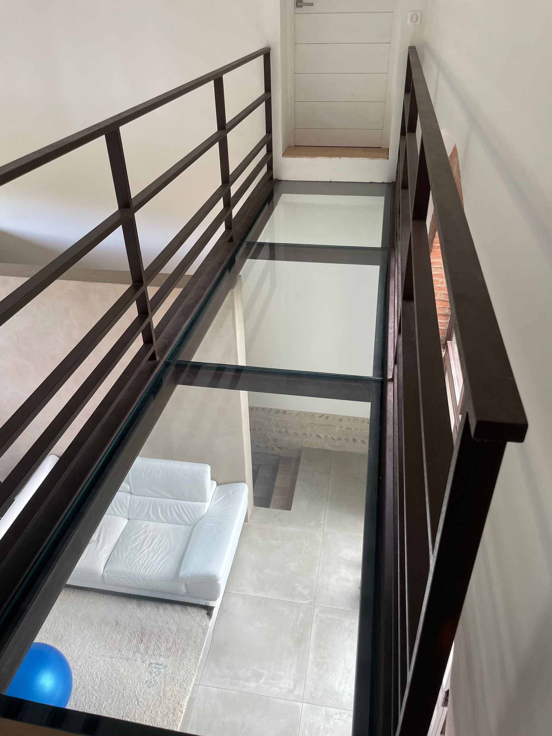 Une passerelle étroite au sol de verre et aux balustrades sombres surplombe un espace de vie avec un canapé blanc et un sol carrelé en contrebas.