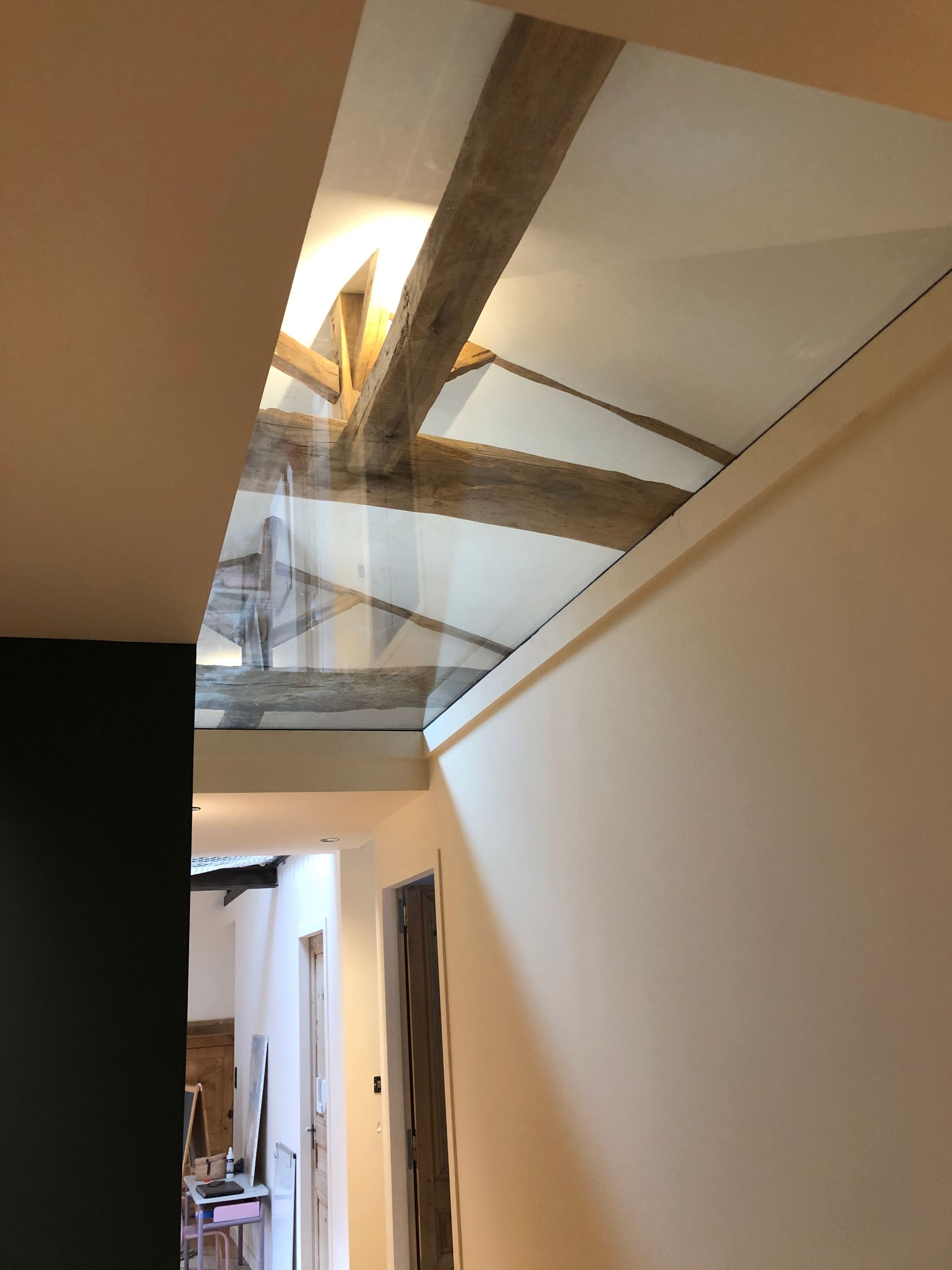 Une section rectangulaire de plafond en verre révèle des poutres en bois rustiques et une charpente blanche au-dessus d'un couloir étroit.