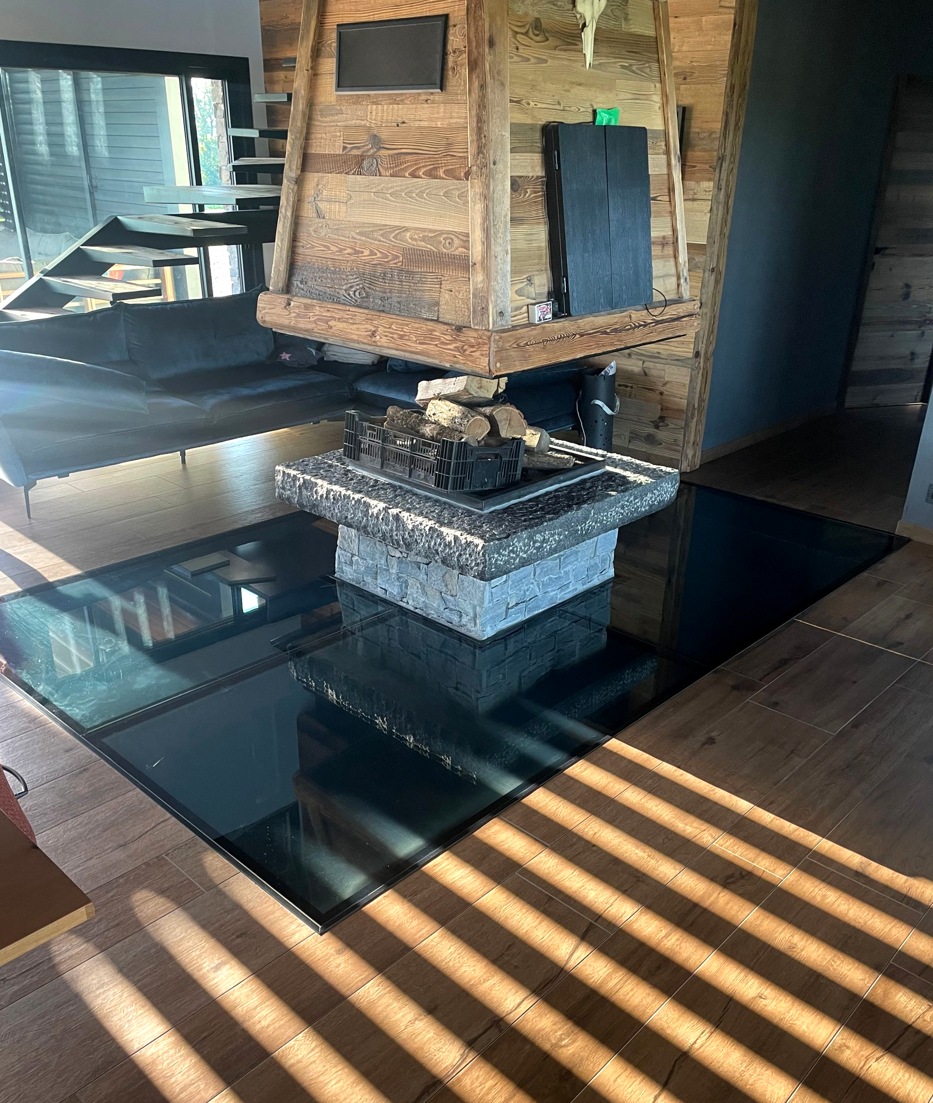 Une cheminée en pierre surplombe un panneau de sol sombre et réfléchissant, au centre d'une pièce au parquet clair veiné.