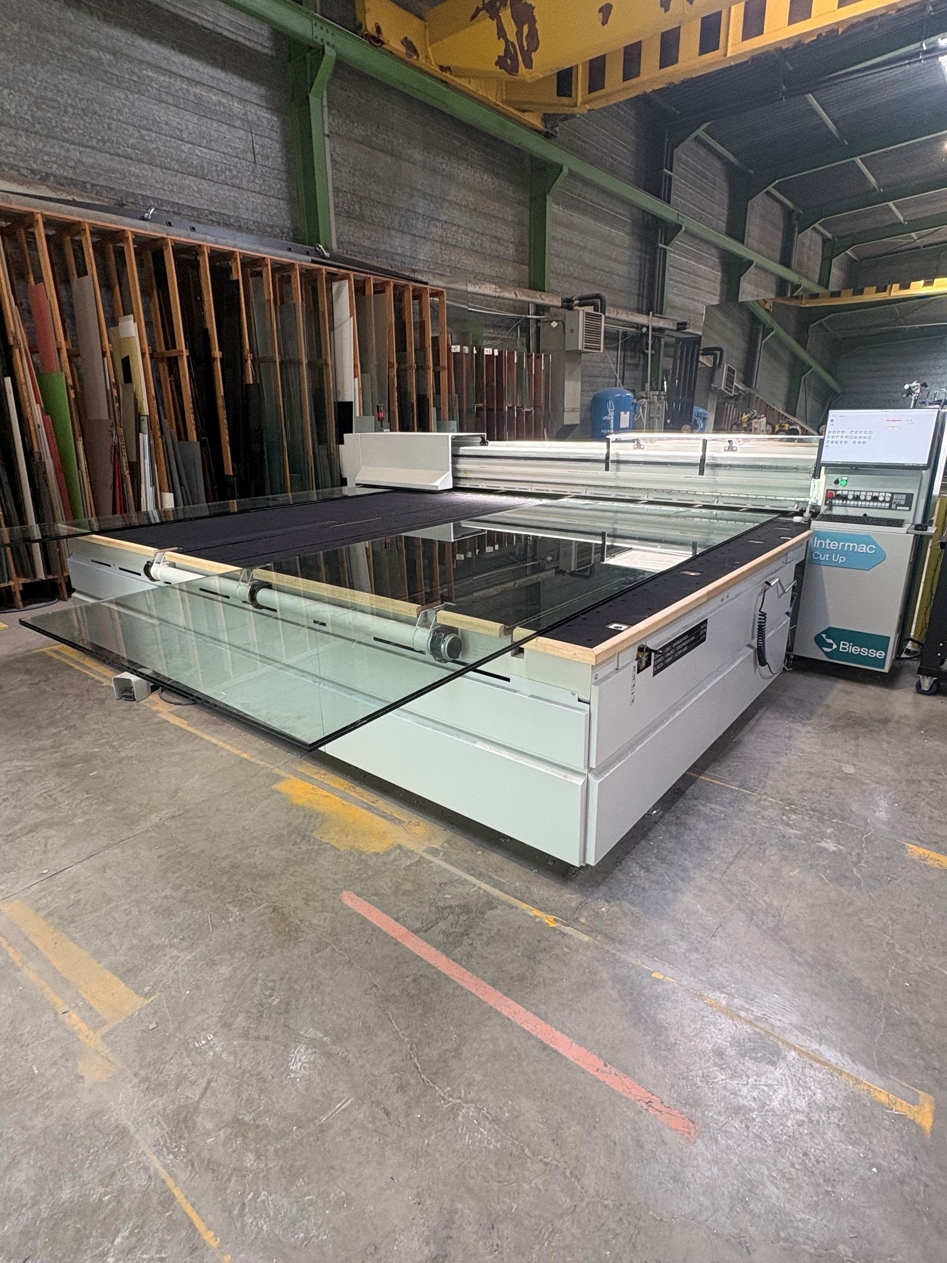 Grande machine de découpe laser industrielle à plat dans un atelier d'entrepôt, avec des matériaux empilés et des marquages ​​au sol en béton.