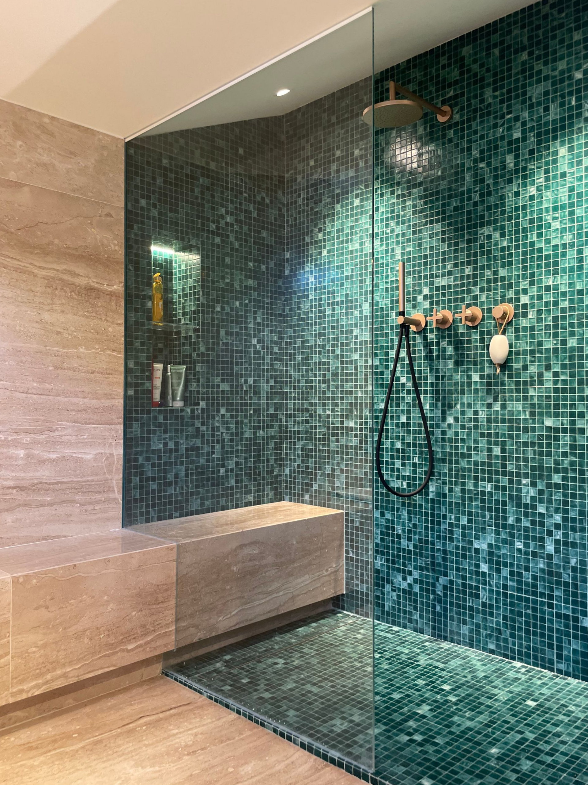 Une douche moderne avec des murs en mosaïque turquoise, une porte en verre, un banc et des accessoires dorés.