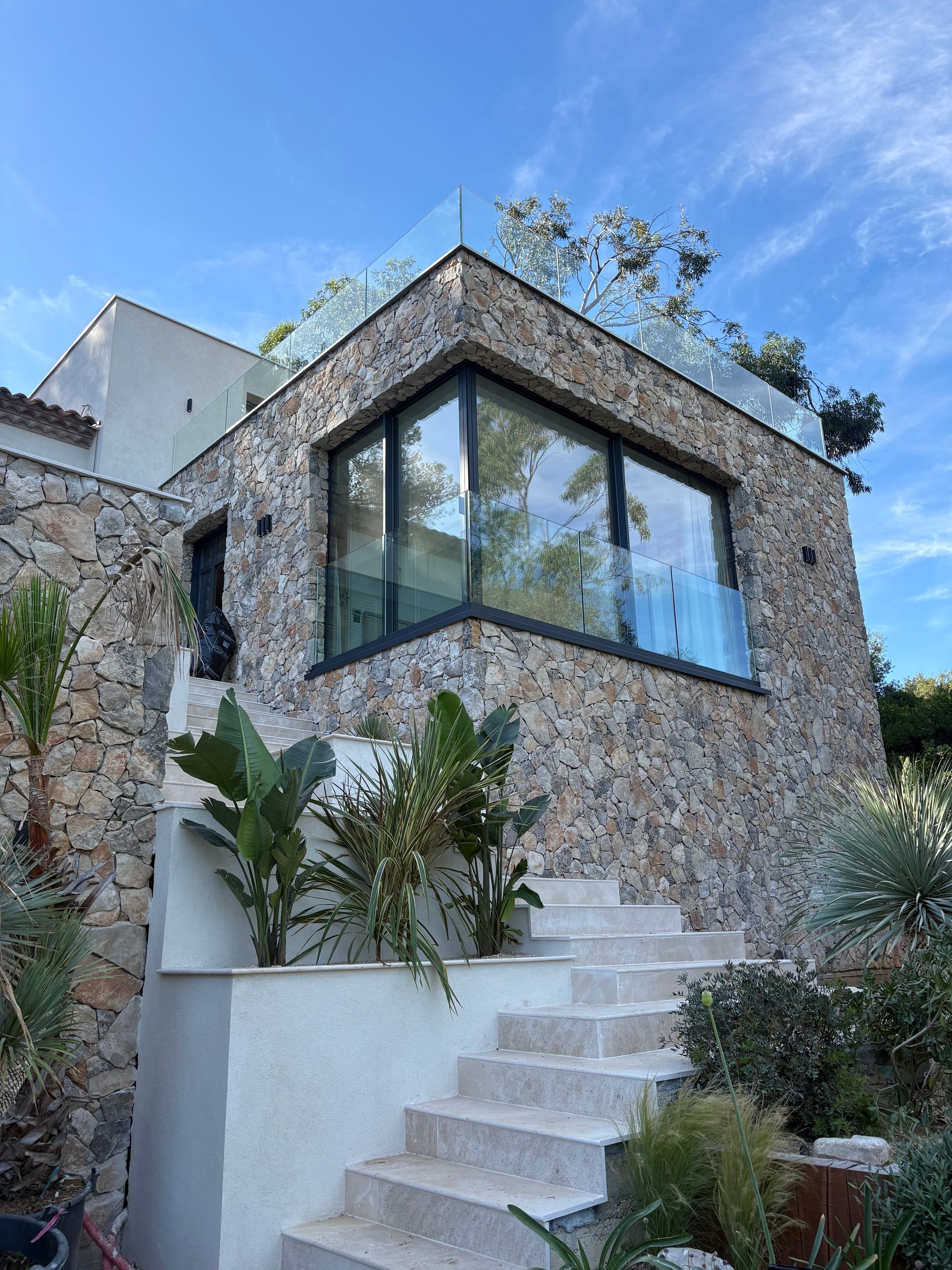 Maison en pierre avec une grande baie vitrée, un escalier blanc et des plantes tropicales sous un ciel bleu.