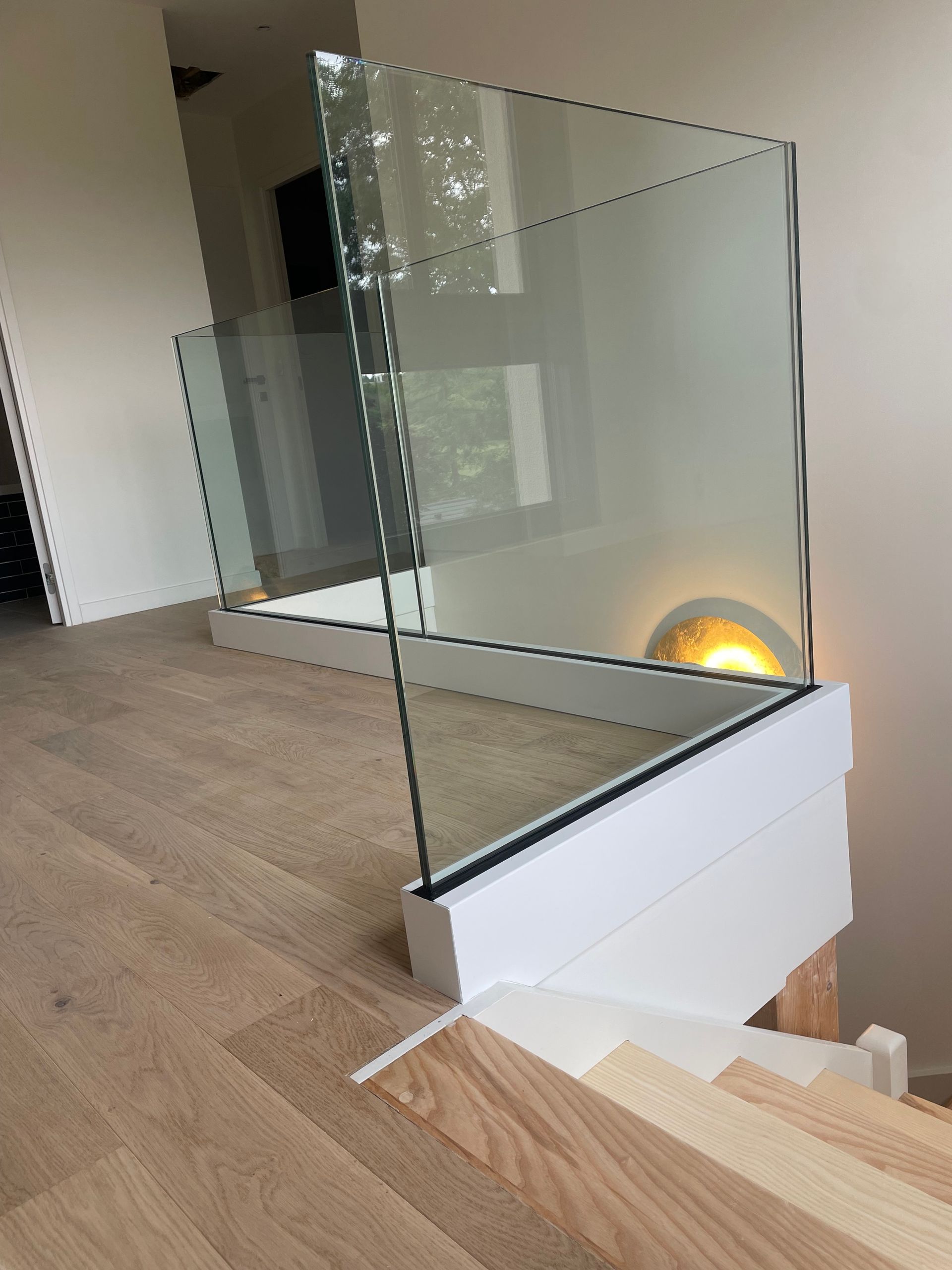 Des panneaux de garde-corps en verre modernes, montés sur un socle en bois blanc, en haut d'un escalier avec un parquet clair.