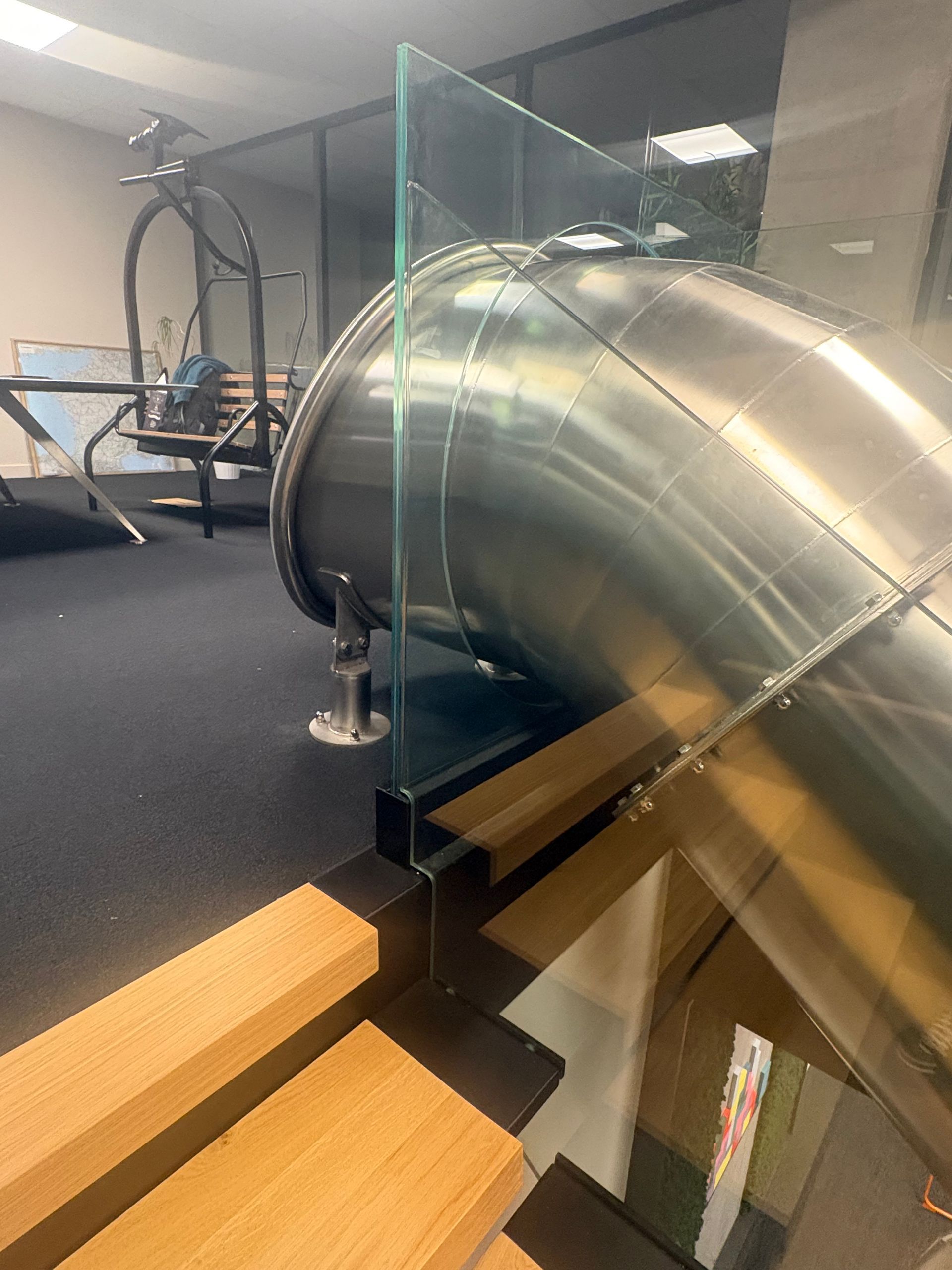 Un toboggan métallique part du bord d'une plateforme de bureau intérieure, sécurisé par une barrière en verre à côté d'un escalier en bois.