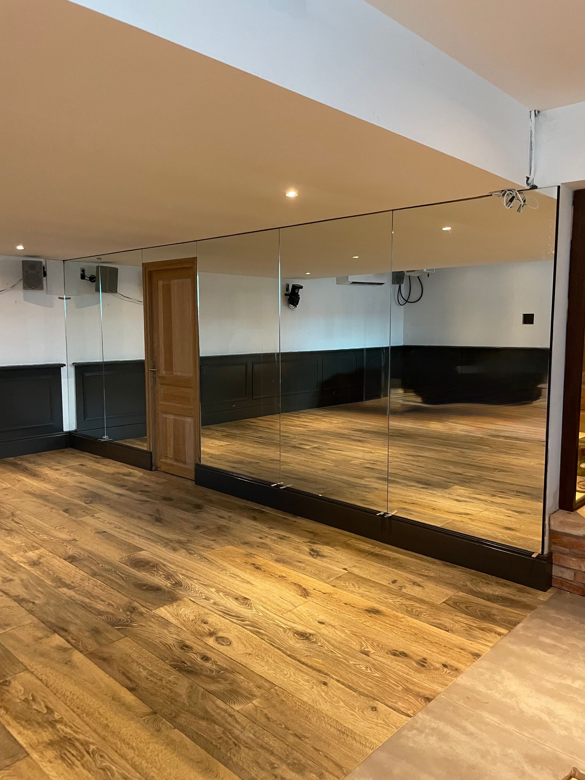 Un mur recouvert de miroirs dans une pièce avec un sol imitation bois, une porte en bois et des plinthes noires.