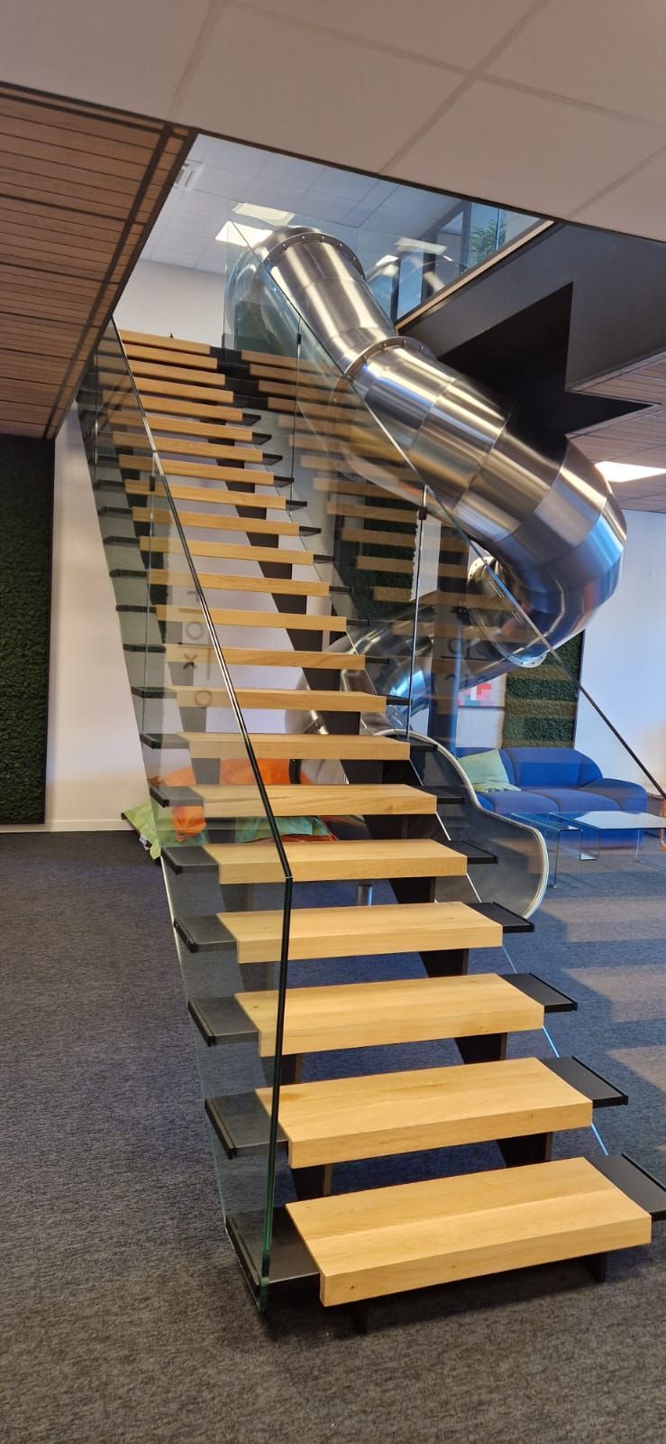 Un escalier intérieur moderne avec des marches en bois et une rampe en verre, accompagné d'un toboggan métallique incurvé.