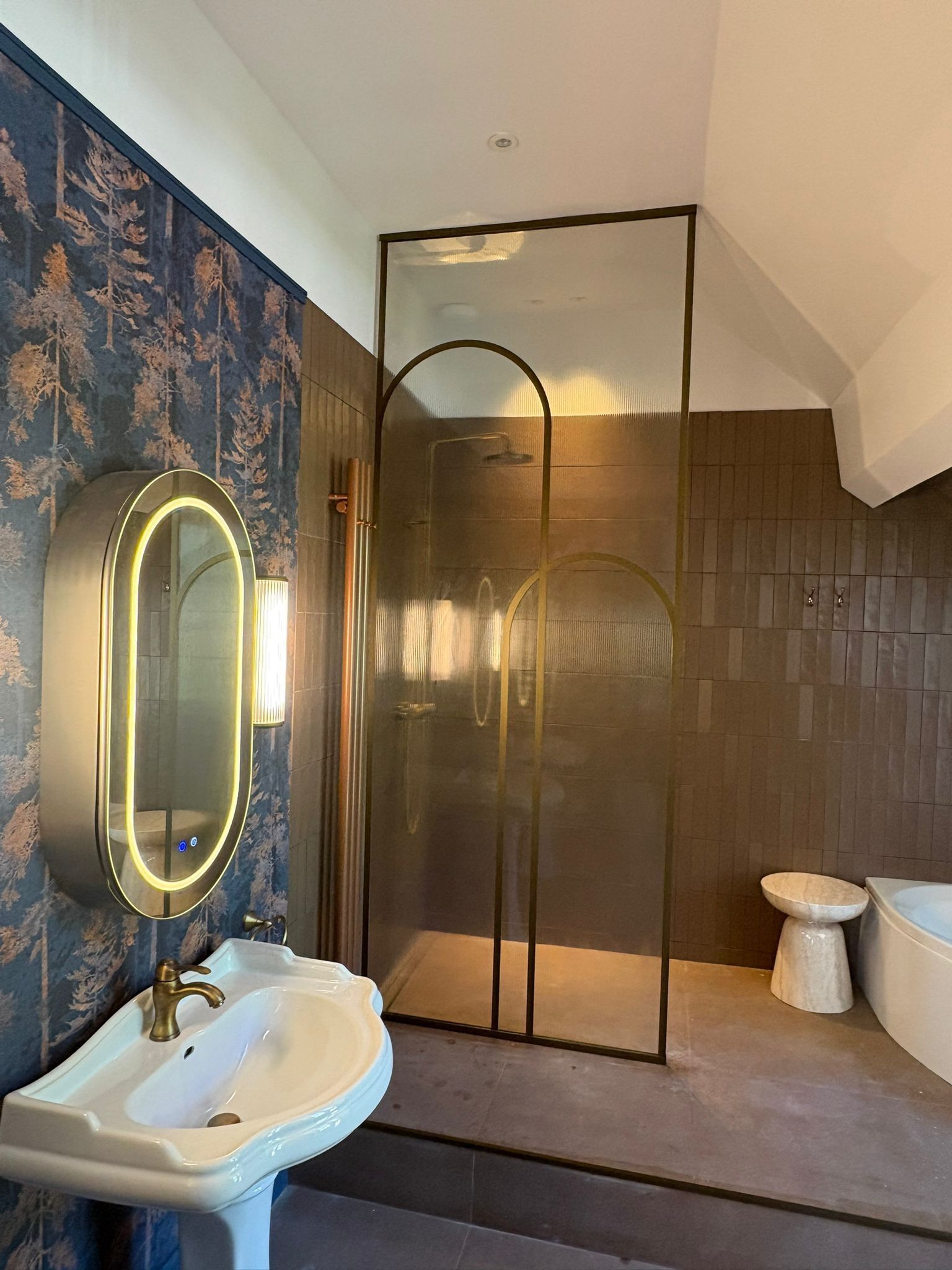 Salle de bains moderne avec douche à l'italienne, lavabo blanc, miroir ovale éclairé et murs carrelés de brun.