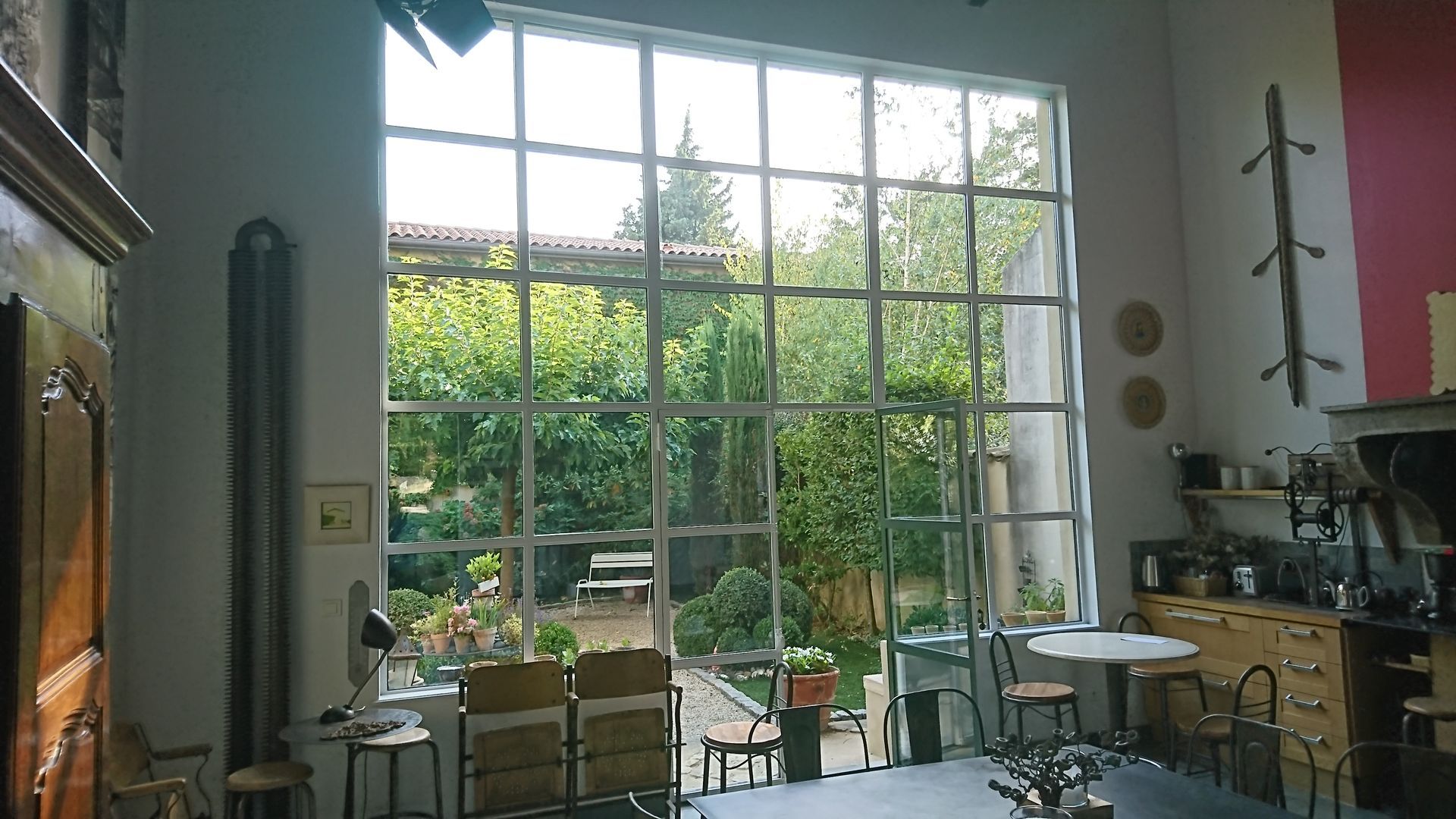 Une grande fenêtre à carreaux donne sur un jardin luxuriant depuis une pièce lumineuse à haut plafond, meublée d'armoires et de meubles.