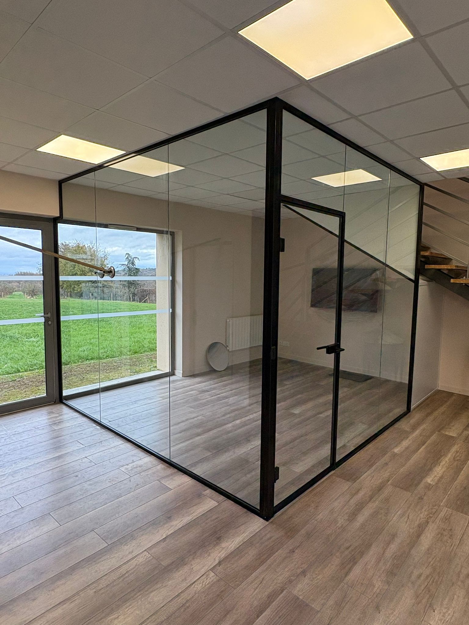 Un espace de bureau moderne caractérisé par une cloison minimaliste en verre aux cadres noirs, contrastant avec une pièce au parquet.