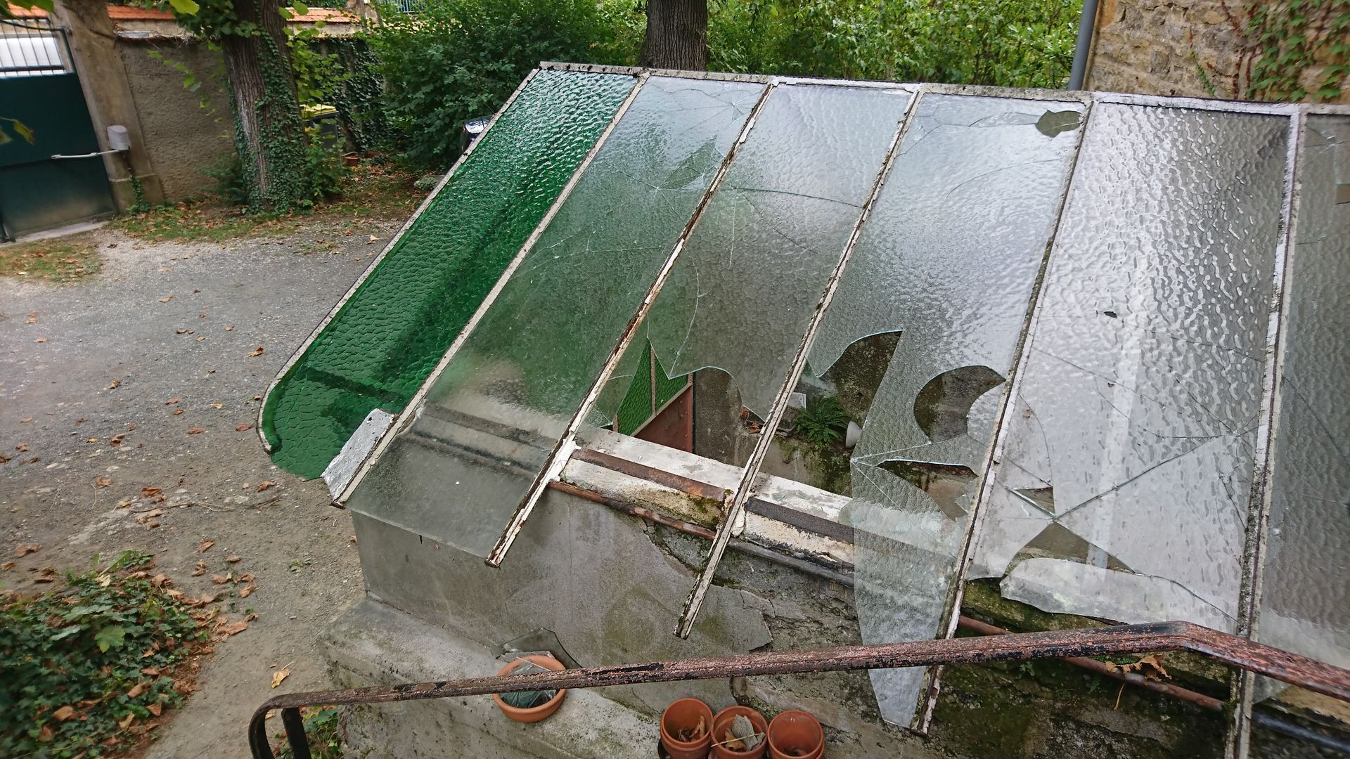 Le toit endommagé d'une serre, composé de plusieurs vitres dont certaines sont cassées, est situé dans un jardin extérieur.
