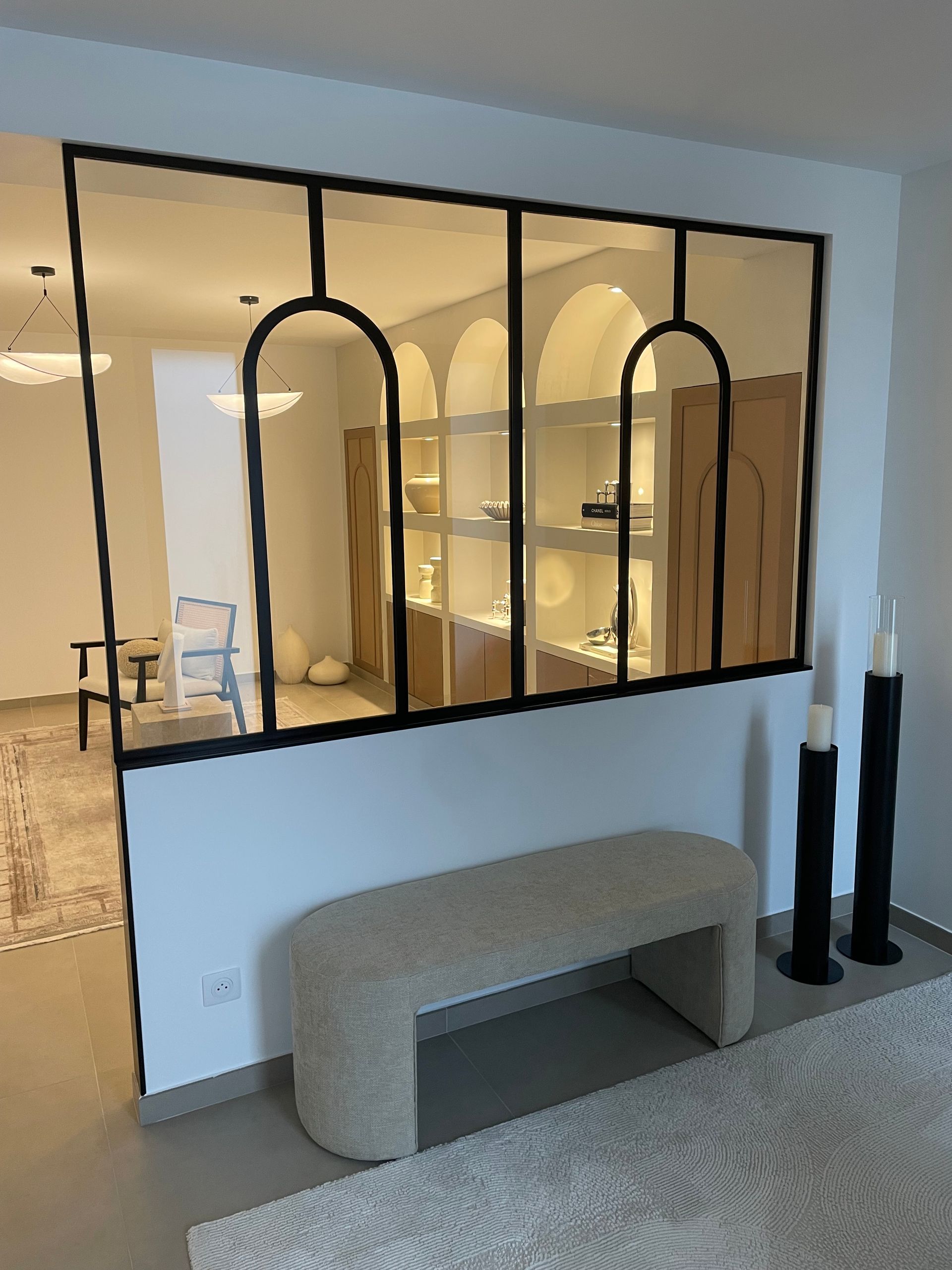 Une cloison moderne avec des cadres arqués en métal noir sur un mur blanc, avec un banc beige devant.