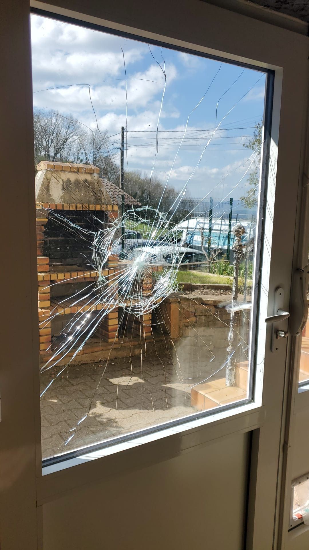 Une porte vitrée présentant un motif de fissures en forme de toile d'araignée, laissant apparaître une cheminée extérieure en briques en arrière-plan.