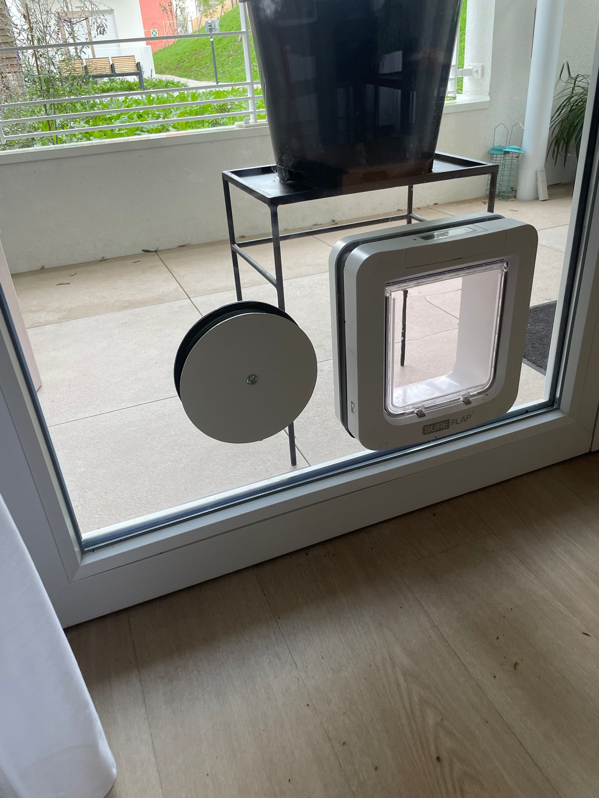 Une chatière et une grille d'aération circulaire sont installées dans une porte-fenêtre vitrée, avec un balcon visible en arrière-plan.