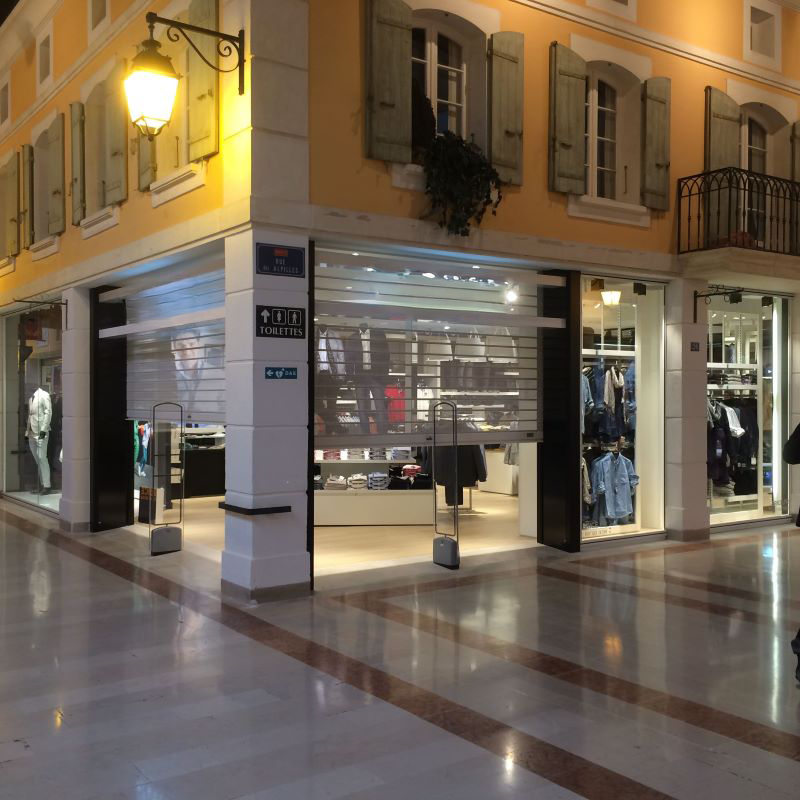 Des boutiques aux volets roulants et des présentoirs de vêtements sont disposés sur une allée claire, sous la façade d'un immeuble à volets.