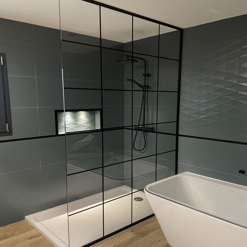 Une salle de bains moderne avec une douche à l'italienne en verre à motif quadrillé, des murs carrelés gris foncé et une baignoire îlot blanche.