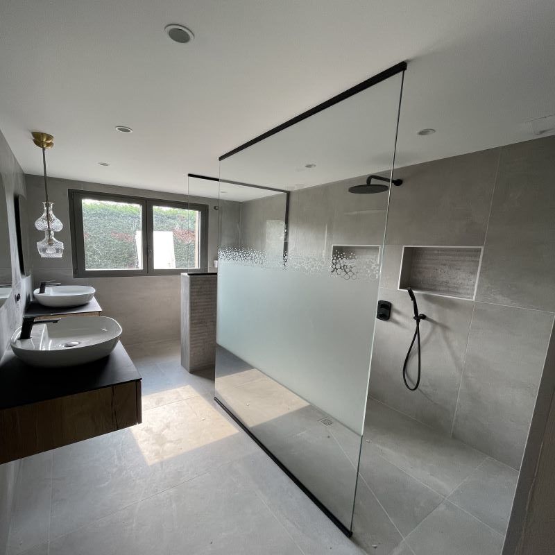Une salle de bains moderne avec un meuble-lavabo suspendu en bois, deux vasques blanches et une douche à l'italienne en verre carrelée de gris.