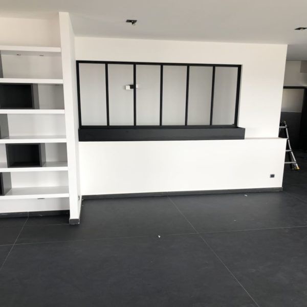 Intérieur avec étagères noires et blanches, fenêtre et sol foncé. Murs blancs.