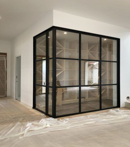 Cave à vin aux parois de verre et à la structure métallique noire, située dans une pièce aux sols en bois clair et aux murs blancs.