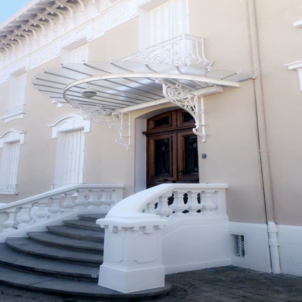Des marches en pierre mènent à une porte d'entrée en bois ornée, surmontée d'un auvent en verre à cadre métallique décoratif.