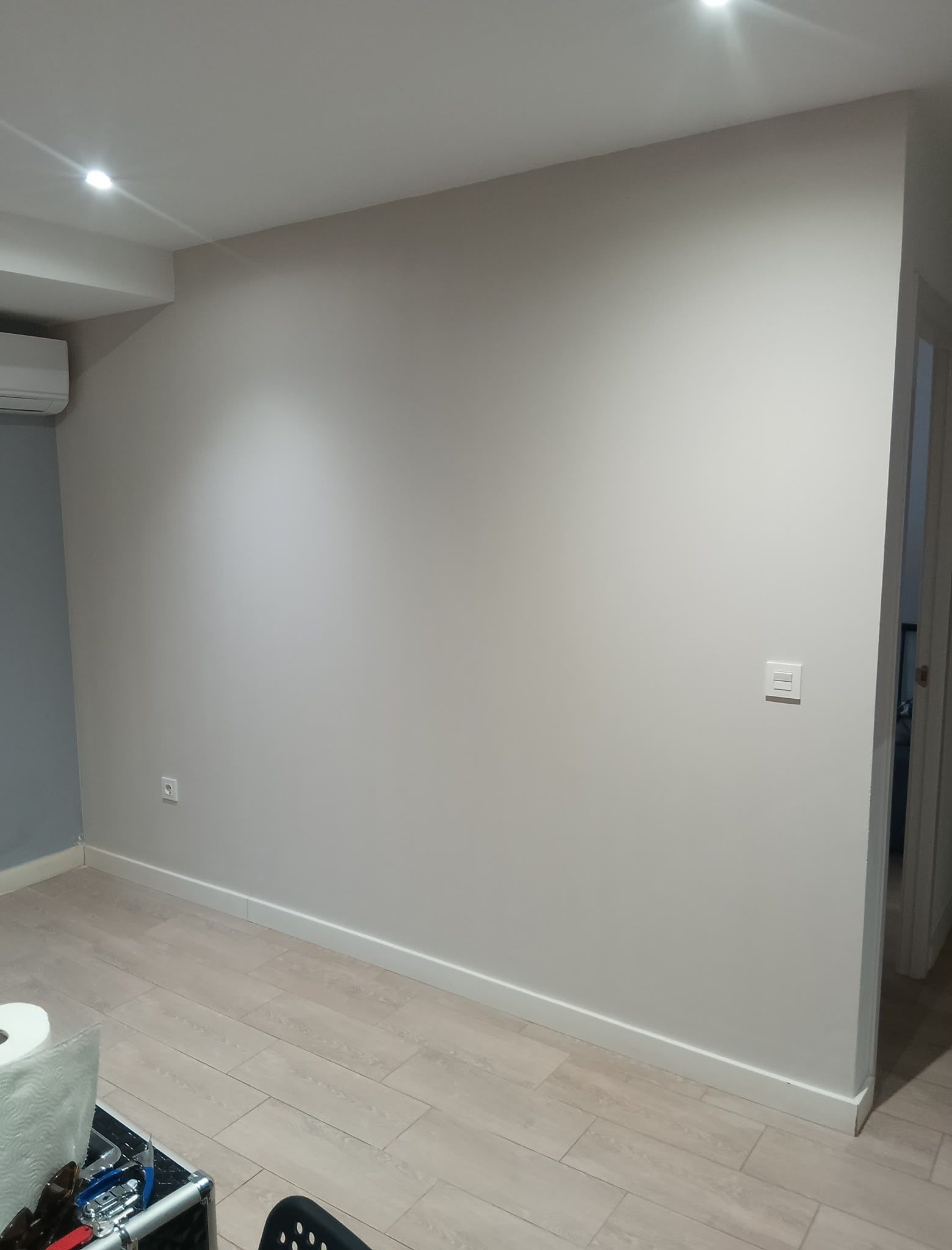 Habitación vacía de color beige con luces blancas en el techo, suelo de madera clara y una puerta a la derecha.
