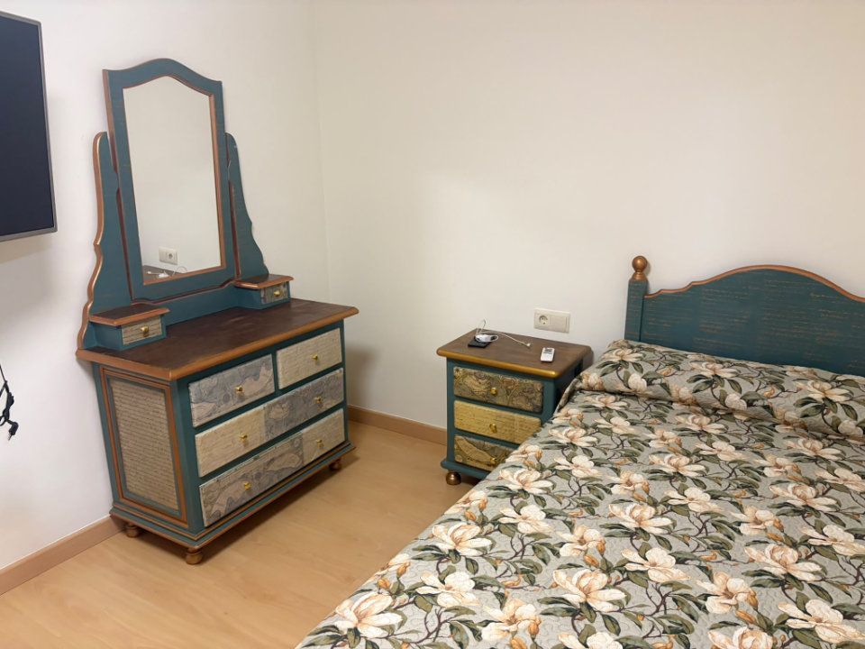 Dormitorio con colcha floral, cómoda azul con espejo y mesita de noche a juego, sobre suelo de madera