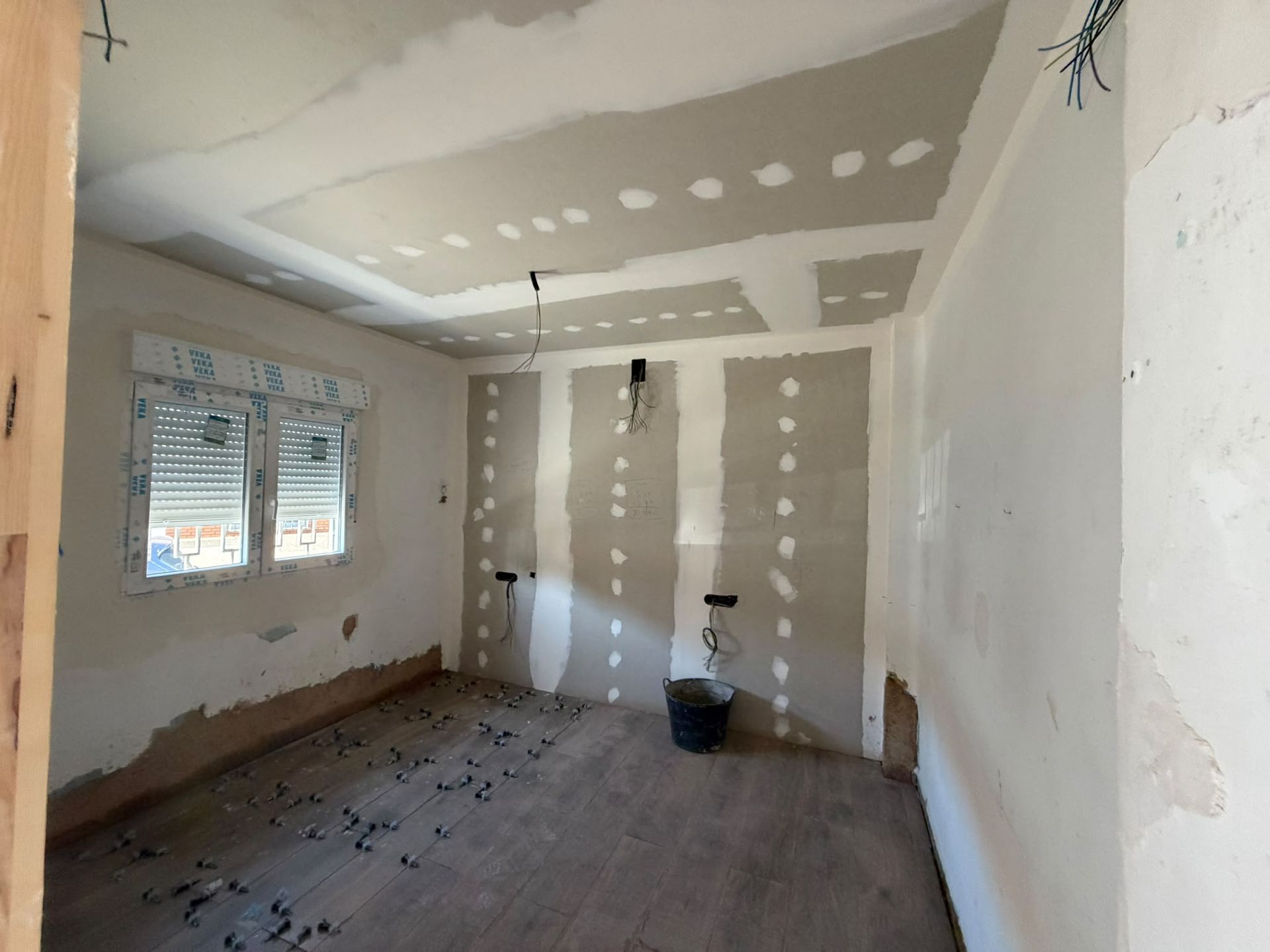 Habitación vacía en obras, con parches de pladur blanco, el suelo al descubierto y una ventana a la izquierda.