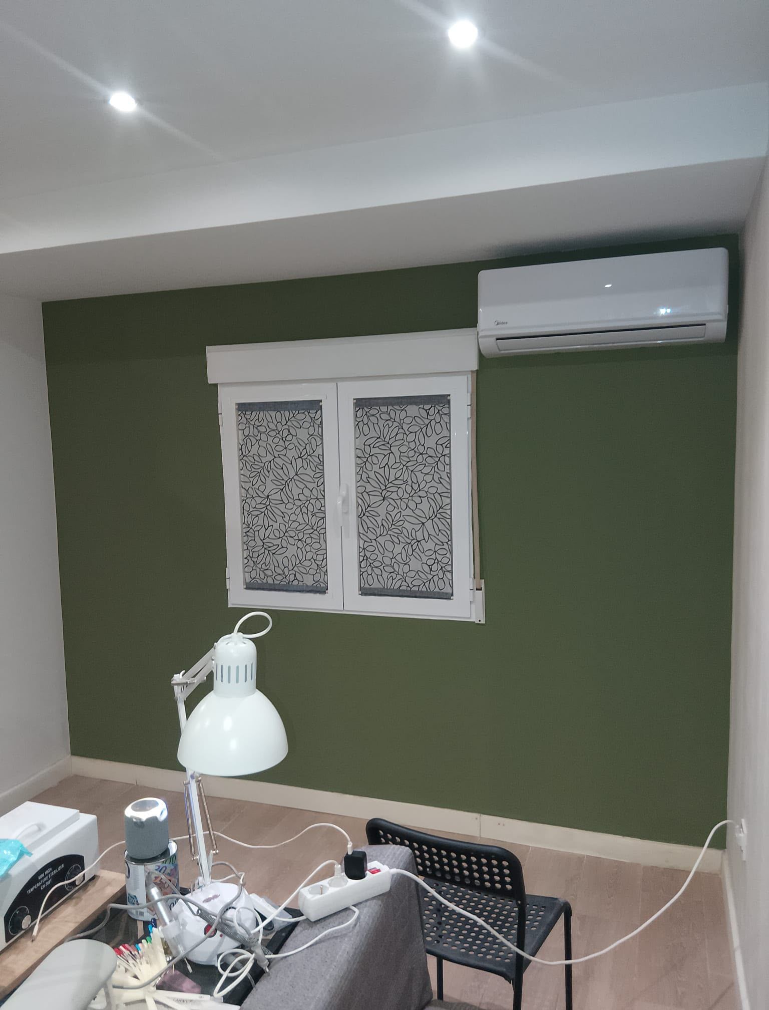 Habitación pequeña con una pared verde, una ventana blanca, un aire acondicionado de pared, una lámpara de escritorio y una silla en primer plano.