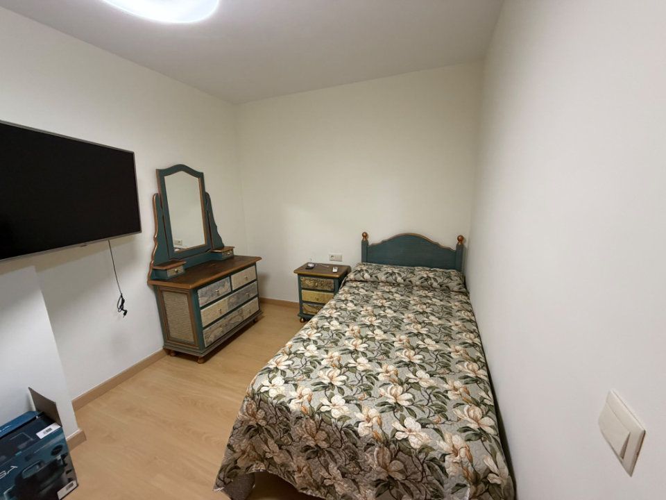Dormitorio pequeño con una cama individual, un televisor montado en la pared, una cómoda y un espejo sobre un suelo de madera clara.