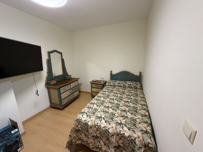 Dormitorio pequeño con una cama individual, un televisor montado en la pared, una cómoda y un espejo sobre un suelo de madera clara.