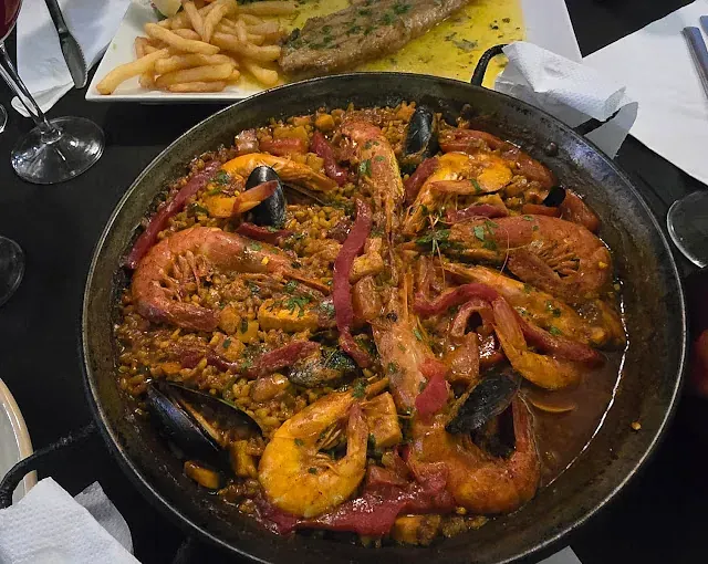 Una sartén con camarones y mejillones reposa sobre una mesa junto a un plato de papas fritas.