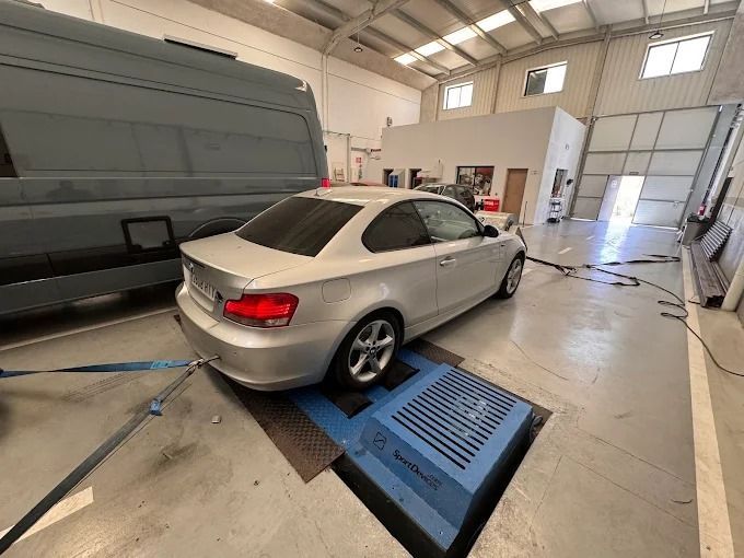 BMW Coupé plateado en un banco de pruebas en un garaje, ruedas traseras sobre rodillos, conectadas con correas y cables.