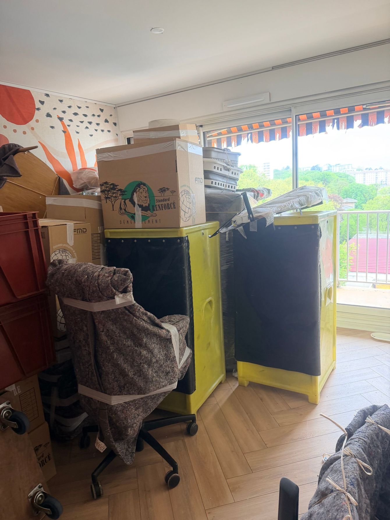 Pièce encombrée de cartons empilés, deux armoires jaunes, une chaise et une fenêtre donnant sur un balcon.