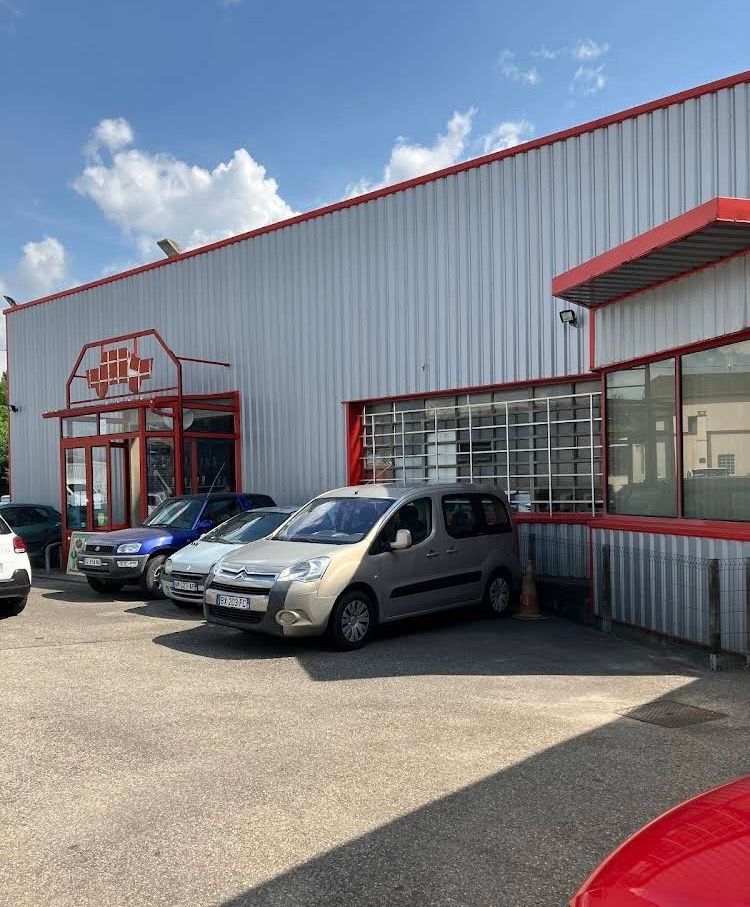 Un garage automobile avec plusieurs véhicules garés à l'extérieur, sous un ciel partiellement nuageux. Le bâtiment est gris et rouge.