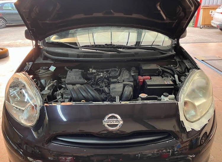 Voiture Nissan noire, capot ouvert, laissant apparaître le compartiment moteur et des optiques en rénovation.