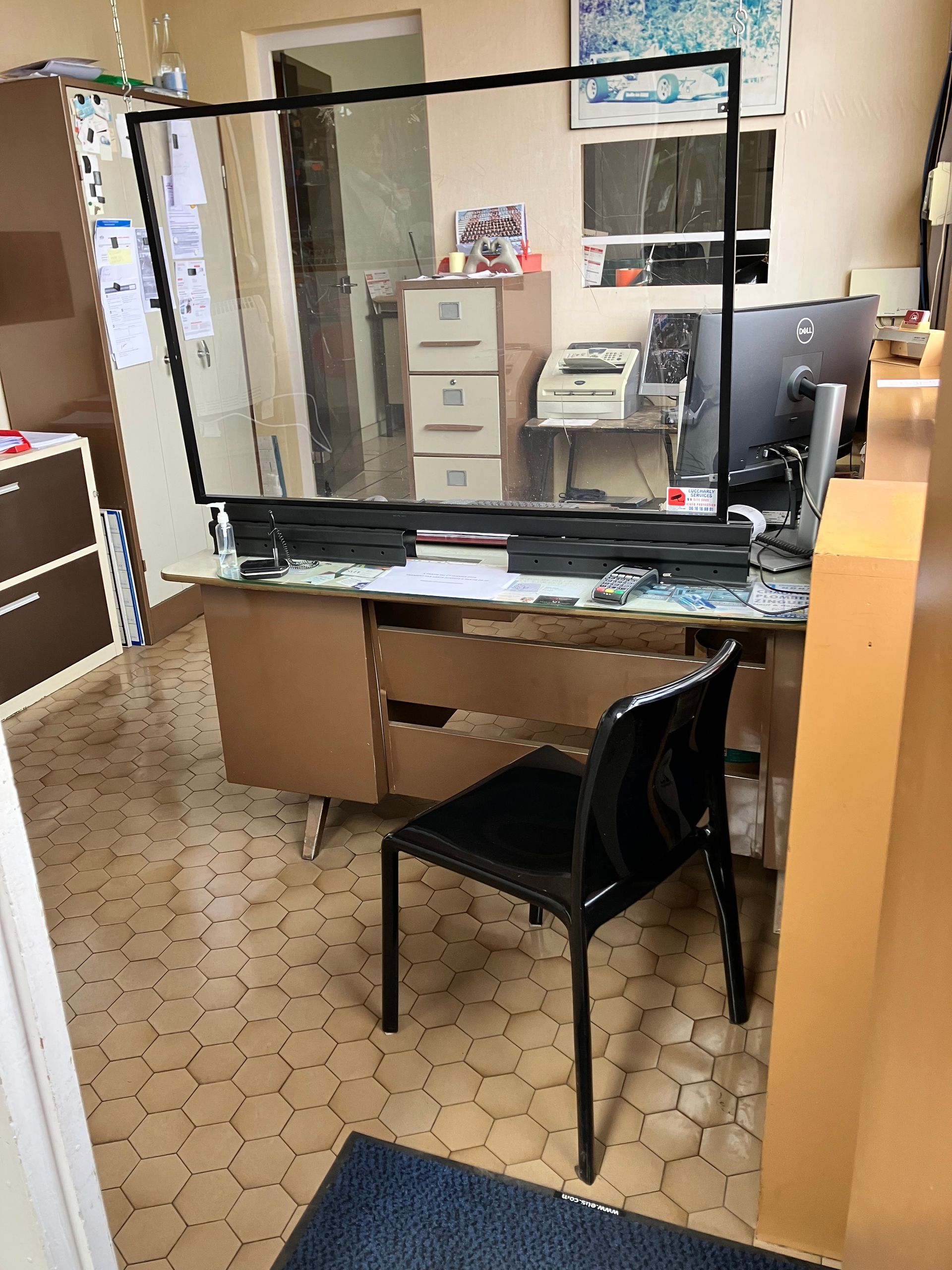 Accueil du garage avec un bureau, une chaise et un grand écran de protection. Un ordinateur, des tiroirs et des documents sont visibles.