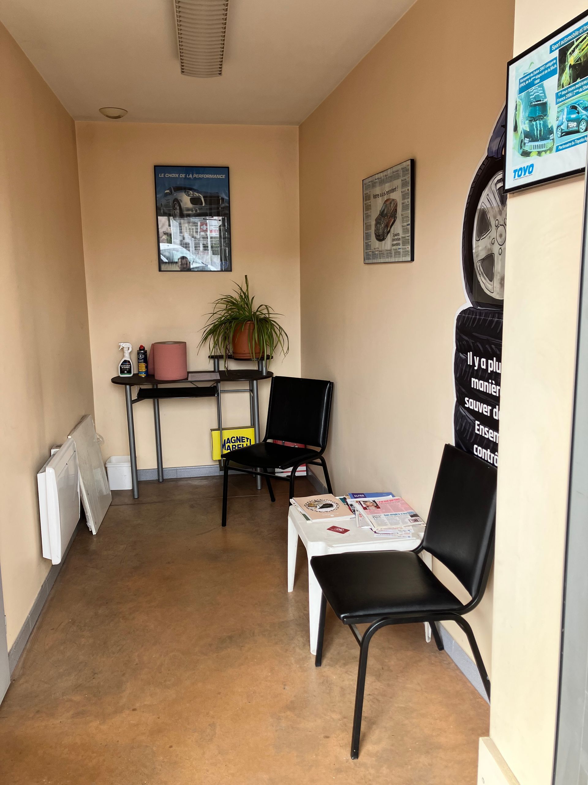 Petit espace d'attente avec des chaises noires, une petite table, des affiches et une plante en pot.