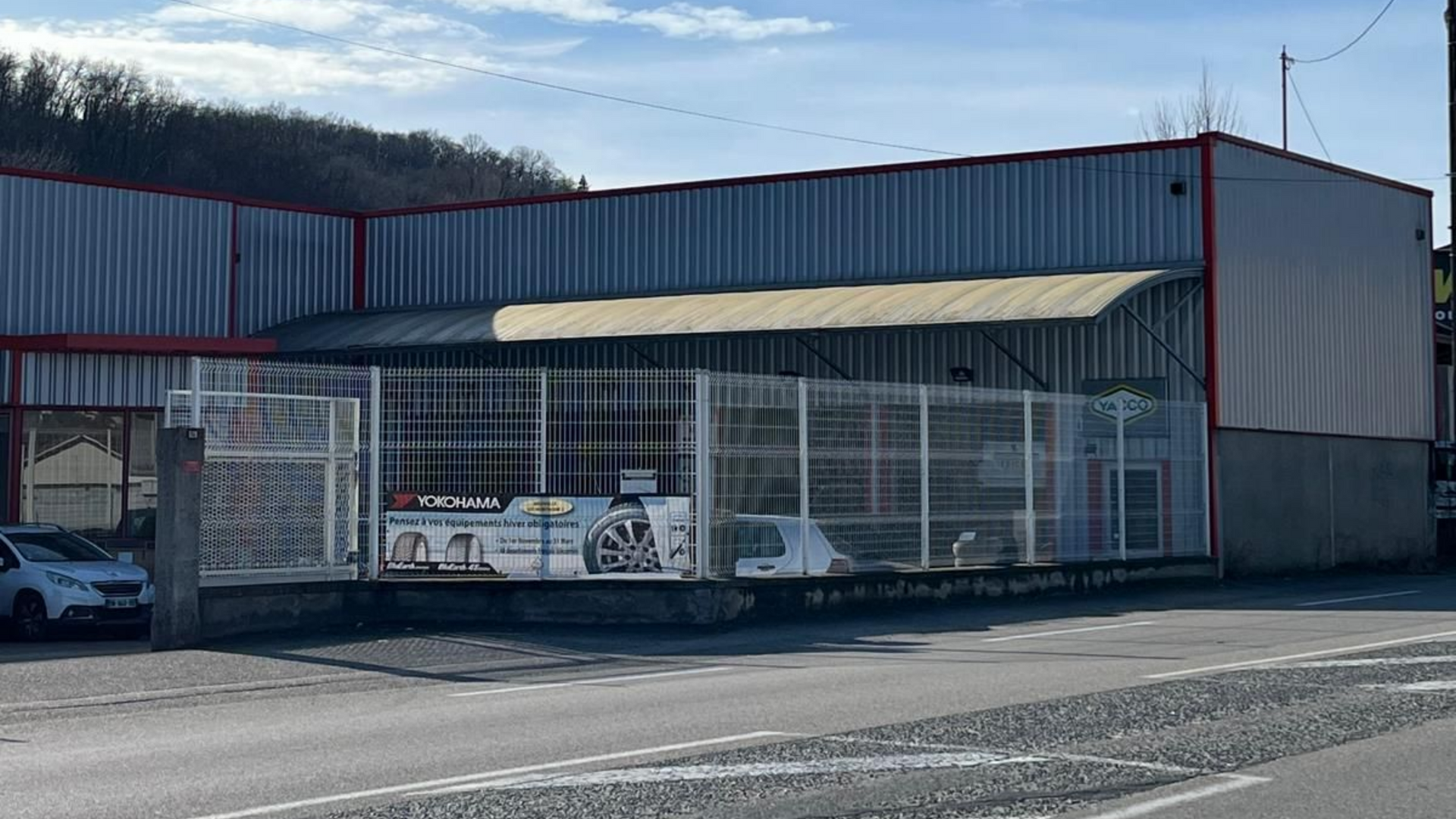 Garage automobile avec bardage métallique gris et rouge, avec une clôture blanche en bordure de rue.