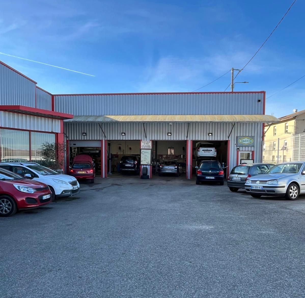 Extérieur d'un garage automobile avec des baies ouvertes et des véhicules à l'intérieur de l'atelier et garés à l'extérieur.
