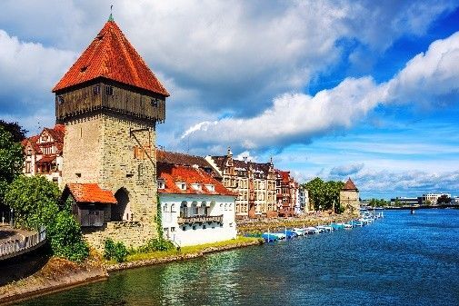 Steinturm an einem Fluss mit roten Dächern und blauem Himmel, neben Gebäuden und Booten am Wasser.