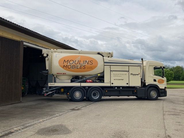 Un camion Moulins Mobiles blanc et jaune est garé à côté d'une grange sous un ciel nuageux.