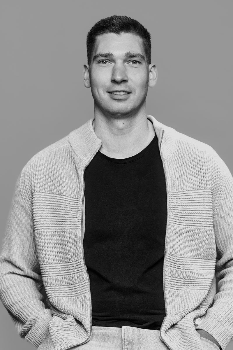 Ein Mann in einem beigen Pullover lächelt vor einem neutralen Hintergrund.