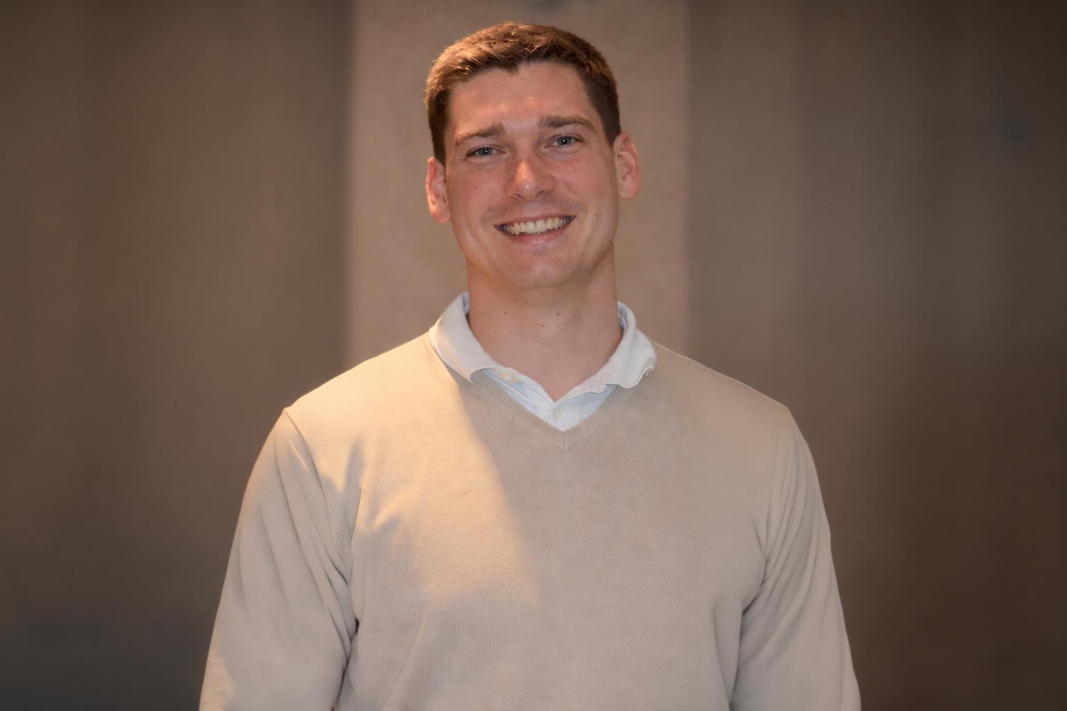 Ein Mann in einem beigen Pullover lächelt vor einem neutralen Hintergrund.