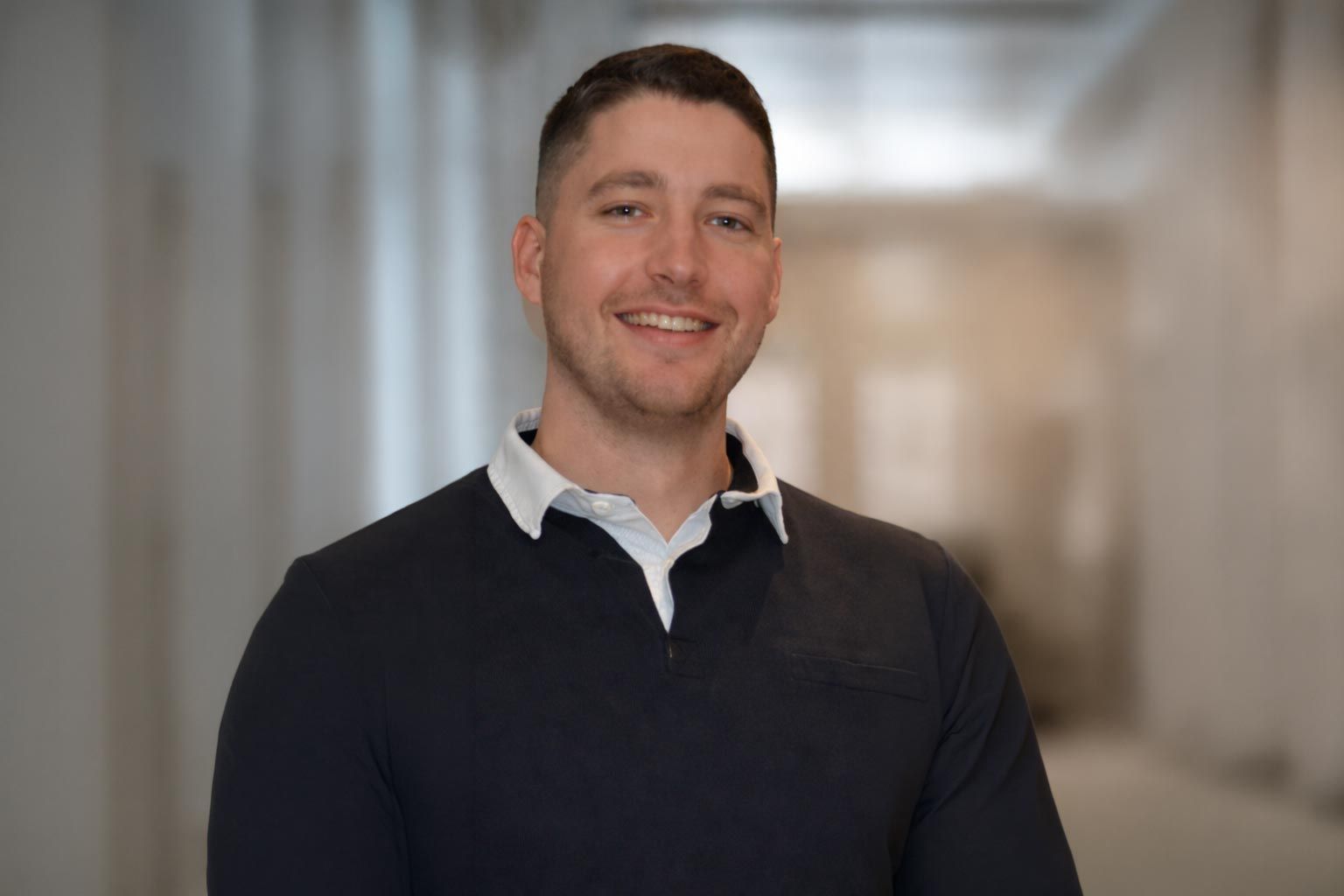 Mann lächelt in blauem Pullover und weißem Kragen. Verschwommener Hintergrund: Flur.