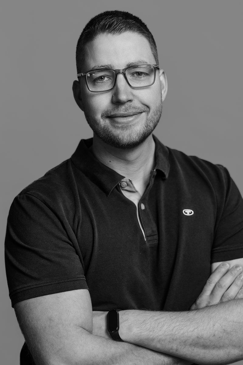 Mann lächelt in blauem Pullover und weißem Kragen. Verschwommener Hintergrund: Flur.
