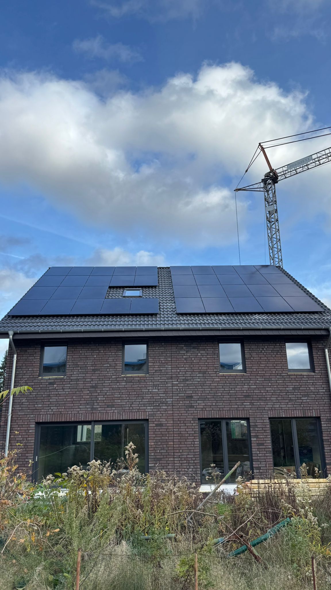 Backsteinhaus mit Solarpaneelen auf dem Dach, Kran darüber, vor bewölktem Himmel.