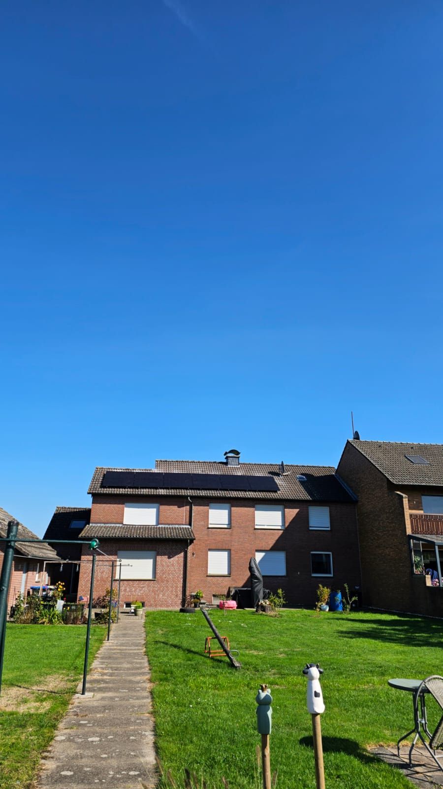 Backsteinhaus mit Solaranlage auf dem Dach unter blauem Himmel. Grüner Rasen und Gehweg davor.