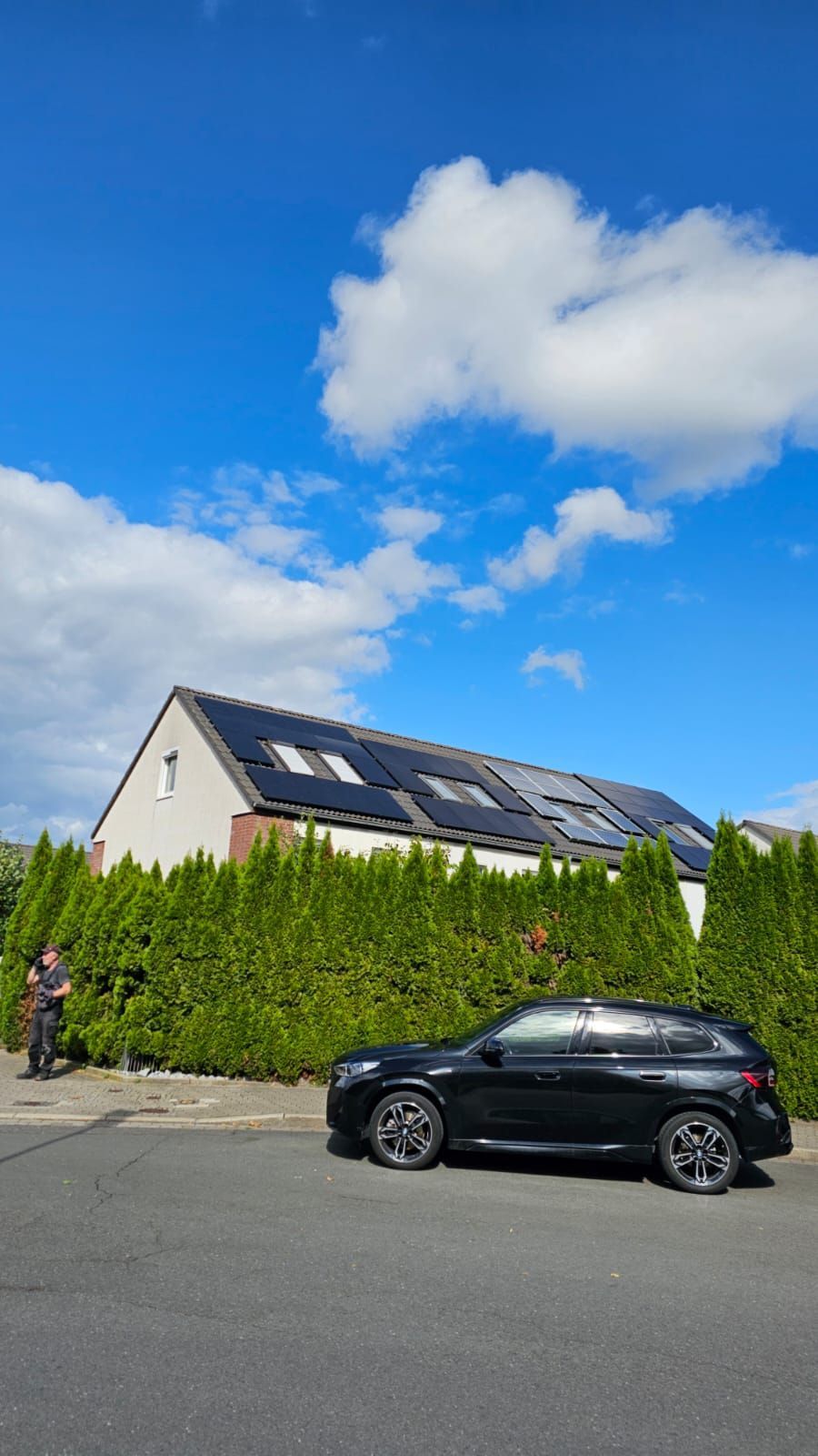 Ein schwarzes Auto parkt vor einem Haus mit Solaranlage auf dem Dach, davor eine grüne Hecke. Eine Person steht daneben. Blauer Himmel.