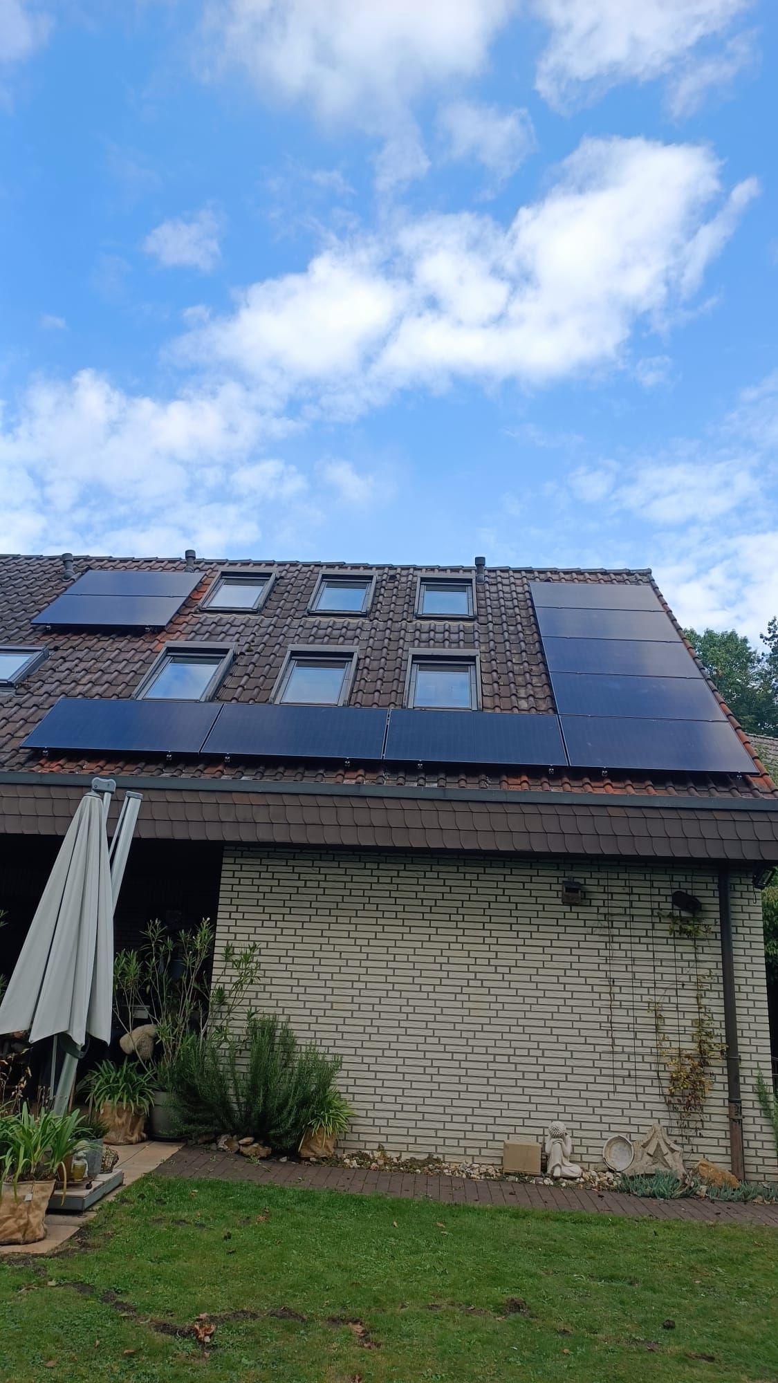 Solarpaneele auf einem Ziegeldach mit Oberlichtern vor einem bewölkten blauen Himmel. Darunter ein Backsteingebäude.